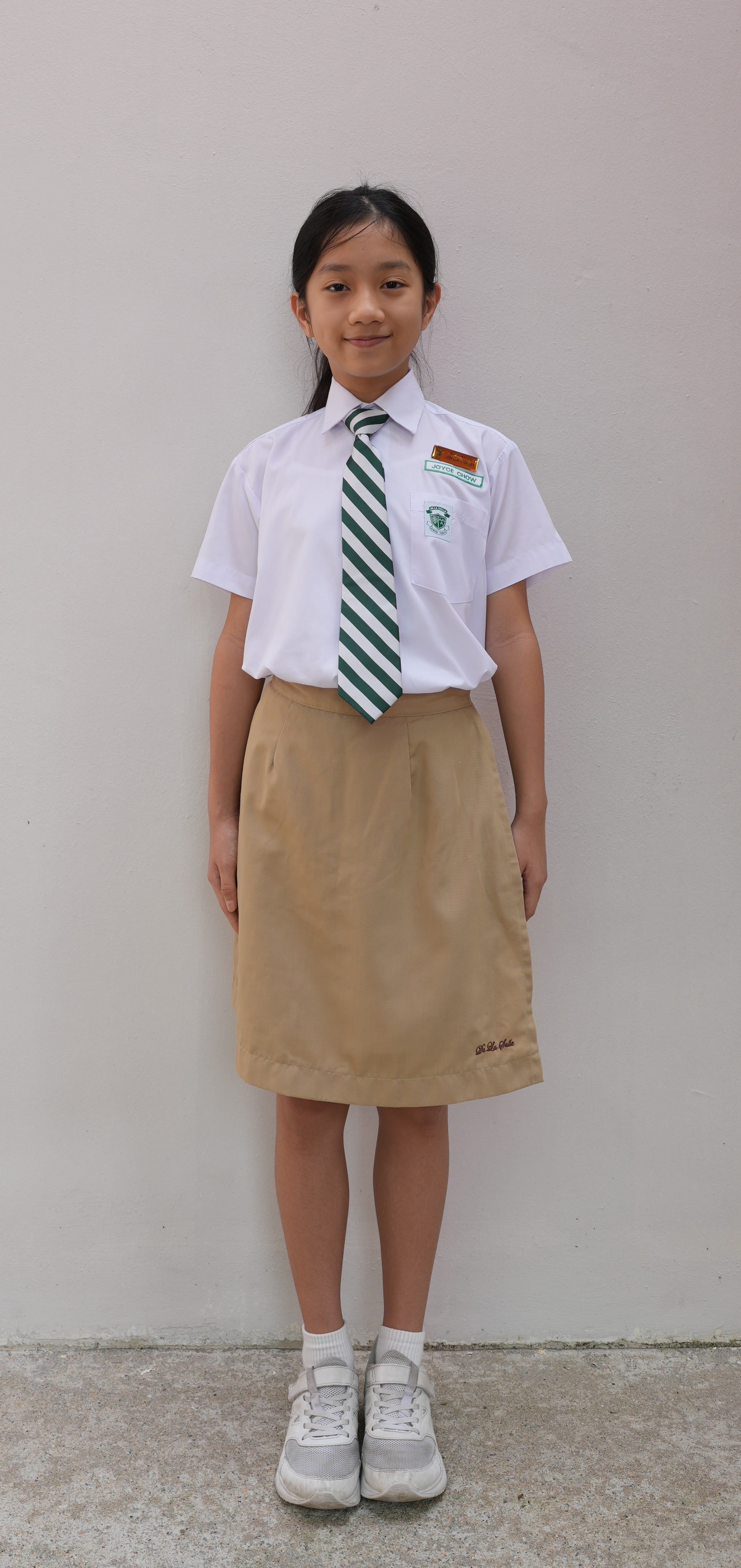 Young girl in white shirt, dark tie, and khaki skirt school uniform standing before green leafy plants.